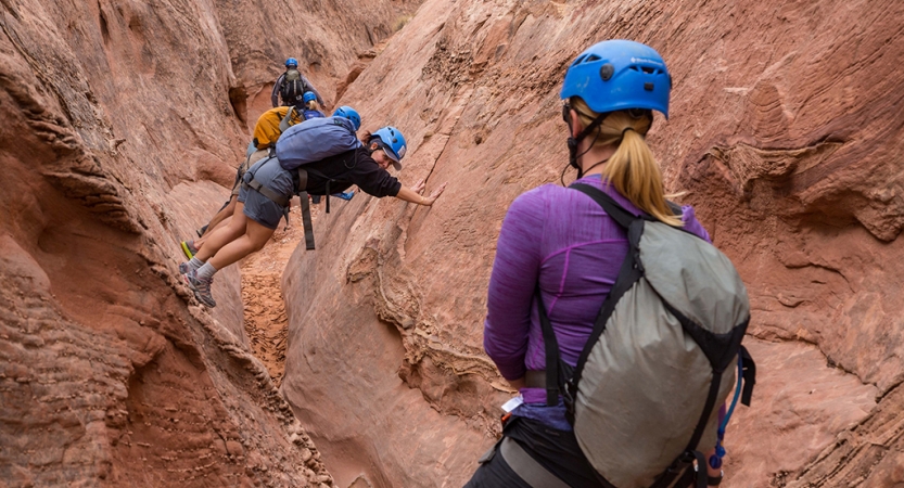 In the foreground, a person looks on as others make their way through a narrow canyon by bracing their hands on one side and their feet on the other. Everyone is wearing helmets.